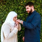 Camila Oliveira Nunes beijando a aliança de seu marido, em sua mão. Ele à olha com um sorriso. Fundo são plantas e ela veste hijad e roupas claras e ele camisa azul.