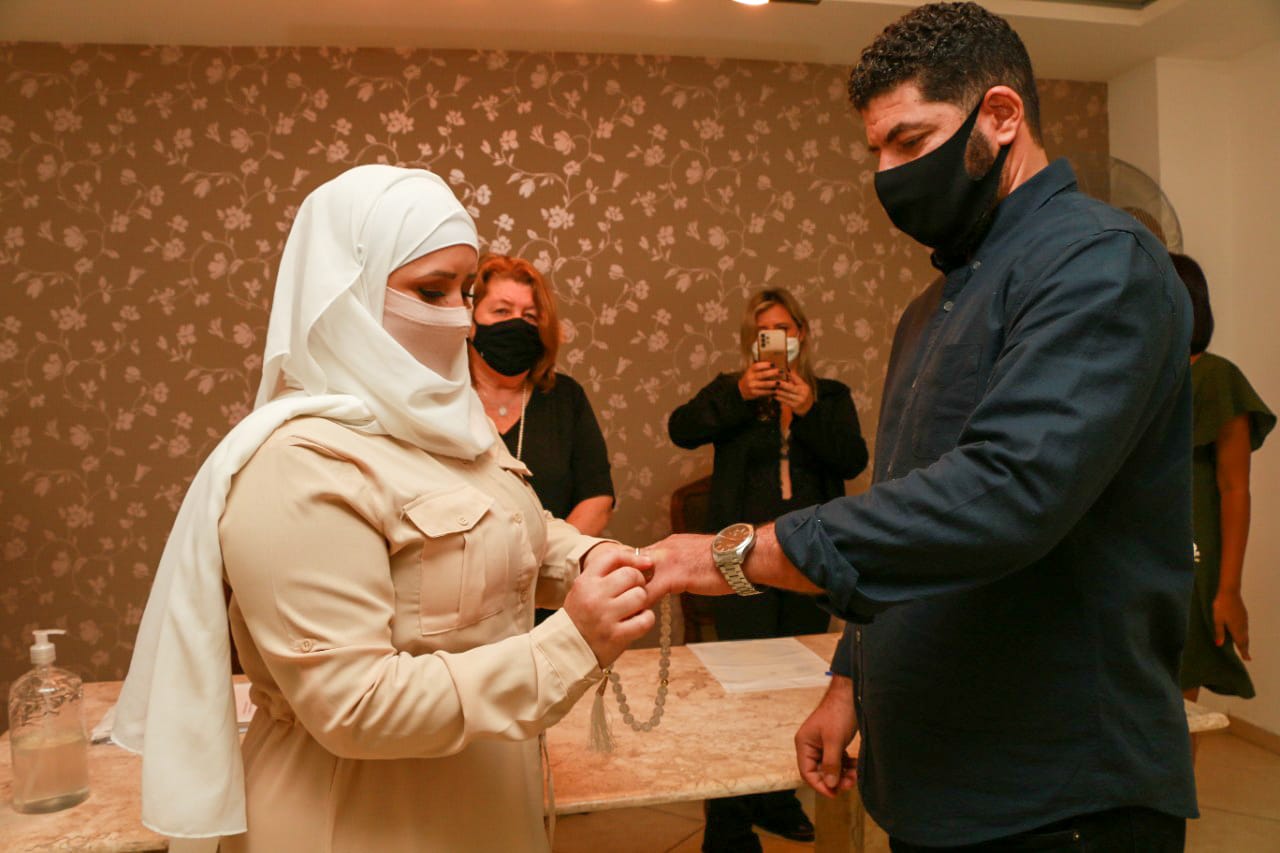 Camila Oliveira Nunes está colocando a aliança em seu marido. Algumas pessoas ao fundo assistindo e fotografando. Ambiente interno do local. Eles estão em primeiro plano, ela utilizando hijab e vestes claras e ele uma camisa escura.